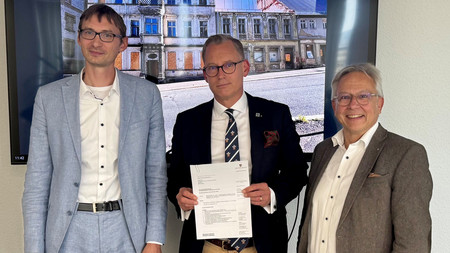 Drei Herren vor einem Bildschirm, auf welchem verfallene Häuser zu sehen sind. Der Mann in der Mitte hält ein Schriftstück in der Hand und zeigt dies in die Kamera